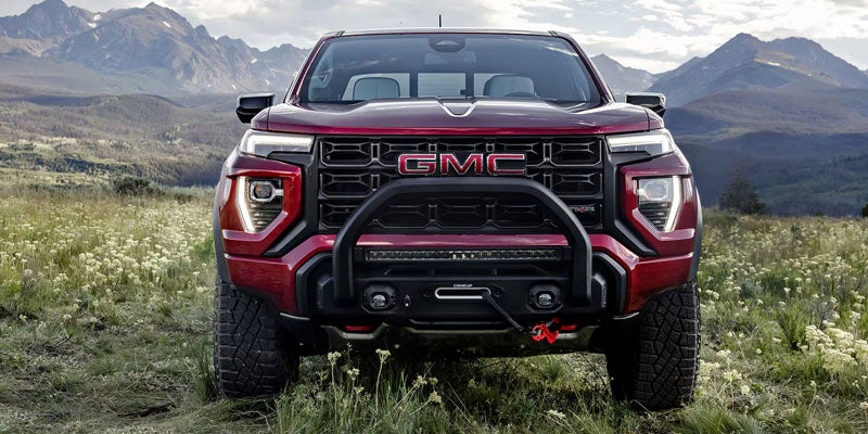 Front view of a red GMC Canyon pickup truck with an off-road grille guard parked in a scenic mountain landscape.